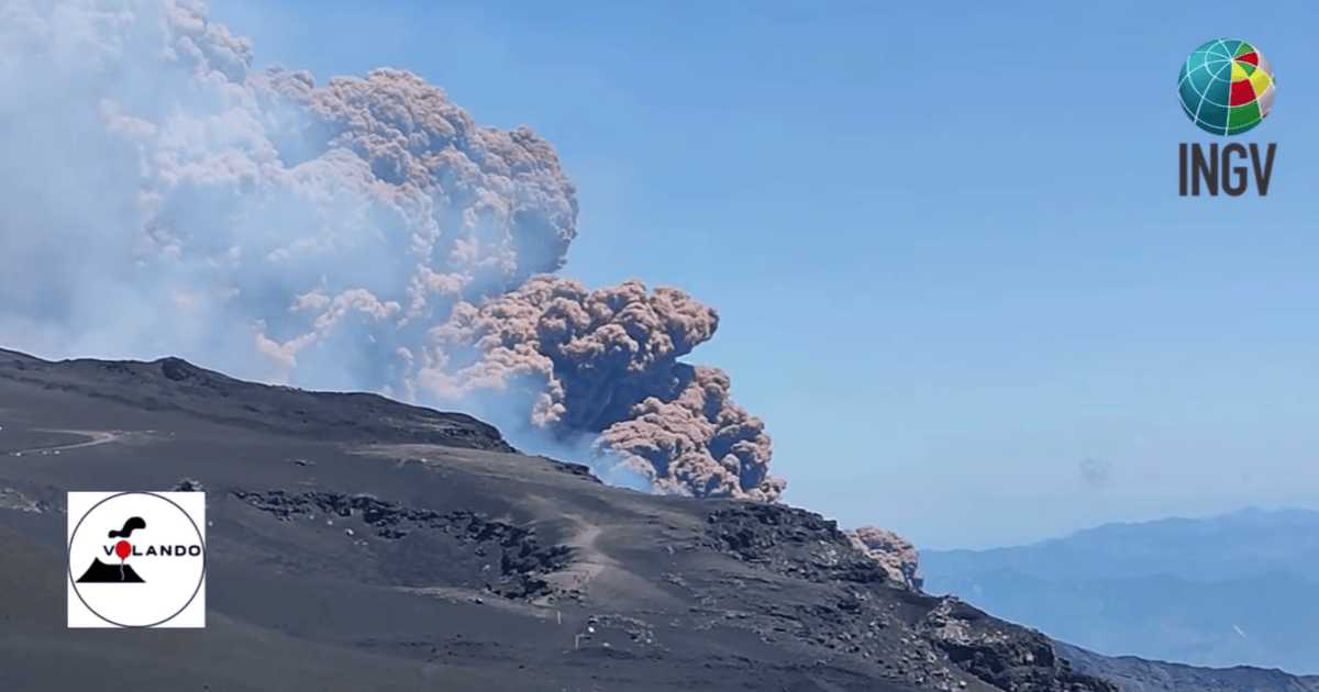 Ash plume rising from Mount Etna’s crater against a clear blue sky, with dark volcanic slopes in the foreground, the INGV logo in the top right, and the Volando logo in the bottom left. 